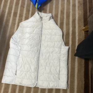 Vit quiltad dunväst Bläck - Snygg vit quiltad dunväst med dragkedja framtill och två sidofickor med dragkedja. Perfekt att slänga över en hoodie eller tröja för extra värme. Enkel och stilren design.