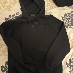 Svart hoodie från Peak Performance - Säljer en svart hoodie från Peak Performance med broderad logga på bröstet. Klassisk modell med huva och magficka, perfekt för en avslappnad stil. Mjuk insida och ribbade muddar vid ärmslut och nederkant.