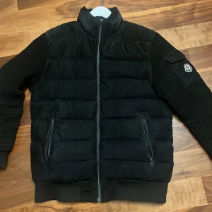 Svart pufferjacka från Moncler - Säljer en svart pufferjacka från Moncler med ribbade ärmar och två dragkedjefickor framtill. Jackan har hög krage och Moncler-logga på ena ärmen. Perfekt för kyliga dagar och riktigt snygg till streetwear.