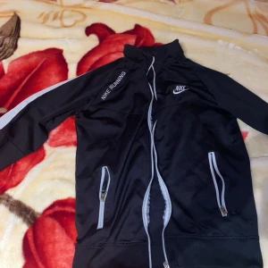 Svart träningsjacka från Nike - Säljer en svart tränings hoodie från Nike med vita detaljer och dragkedja. hoodien har hög krage, två fickor med dragkedja och texten 'NIKE RUNNING' på bröstet. Perfekt för löpning eller sportiga dagar.