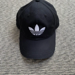 Svart keps från Adidas Originals - Svart keps från Adidas Originals med den klassiska vita trefoil-loggan broderad framtill. Justerbar rem baktill med metallspänne. Kepsen är i bomull och har en böjd skärm. Helt ny, aldrig använd och inga defekter. Pris kan diskuteras vid snabb affär.