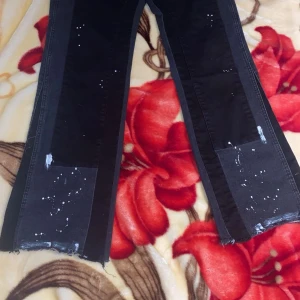 Svarta jeansbyxor med slitna detaljer - Säljer ett par svarta jeansbyxor med breda ben och slitna detaljer nertill. Byxorna har vita färgstänk och patchade partier på framsidan. Perfekta för dig som gillar en edgy och unik stil.