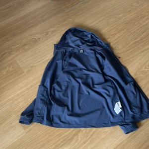 Blå vindjacka från Under Armour - Snygg svart vindjacka från Under Armour med huva och dragkedja. Jackan har en stilren design med Under Armour-logga på bröstet och text på ryggen. Köpt för 1300, mitt pris 1000 pris kan diskuteras.