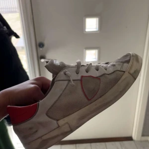Phillipe model skor - Säljer ett par vita sneakers med röd detalj på hälen och sidan. Skorna har snörning och platt sula, samt rund tå. Perfekta för en avslappnad stil och passar till det mesta.