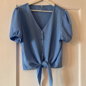 Blå knytblus från JDY - Säljer en ljusblå blus från JDY med v-ringning, knappar framtill och knyt i midjan. Blusen har korta puffärmar med resår och är tillverkad i ett lätt, strukturerat tyg. Perfekt för vår och sommar!