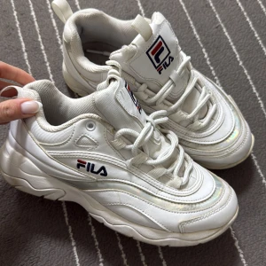 Vita sneakers från Fila - Säljer ett par vita Fila sneakers med chunky sula och snörning. Skorna har Fila-logga på sidan och plösen samt diskreta skimrande detaljer längs sidorna. Perfekta för dig som gillar sportig och trendig stil.