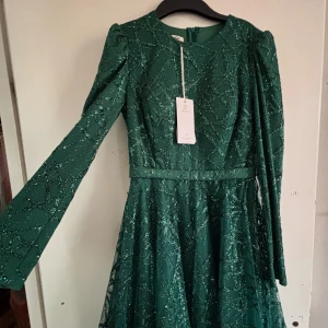 Glittrig grön långklänning med bälte - Säljer en elegant grön långklänning med glittrigt mönster och lång ärm. Klänningen har rund halsringning, markerad midja med bälte och ett skirt yttertyg med glitterdetaljer. Perfekt för speciella tillfällen när du vill sticka ut.