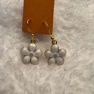 Blommiga örhängen i guld och vitt - Säljer ett par söta örhängen med blommor i vit emalj och guldfärgade detaljer. Blommorna har fem kronblad och små pärlliknande detaljer i mitten. Perfekta för att ge en romantisk touch till din stil. Krokarna är i sterling silver