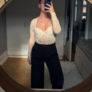 Aprikos mesh body med broderade blommor - Säljer en aprikos mesh body med långa ärmar och vackra broderade blommor. Toppen har en korsettliknande design med markerade kupor och transparent meshmaterial. Perfekt för att skapa en trendig och feminin look.