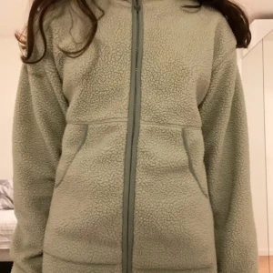 Ljusgrön fleecejacka från Lindex - Mysig ljusgrön fleecejacka från Lindex med dragkedja framtill och två fickor. Jackan har lång ärm och är perfekt för kyliga dagar. Passar barn i storlek 134/140 (8-10 år).