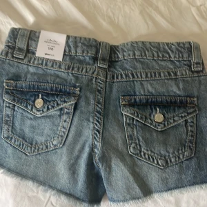 Chunky denim shorts från Gina Tricot - Säljer ett par chunky denim shorts från Gina Tricot i klassisk blå tvätt. Shortsen har låg midja, fransade benslut och slitna detaljer framtill. Bakfickor med lock och knapp. Perfekta för sommaren!