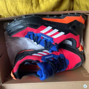 Adidas Terrex AX4 GTX vandringssko - Säljer ett par Adidas Terrex AX4 GTX vandringsskor i rött, blått, svart och orange. Skorna har grov sula för bra grepp, snörning och är märkta med Terrex på sidan. Perfekta för utomhusaktiviteter och äventyr.