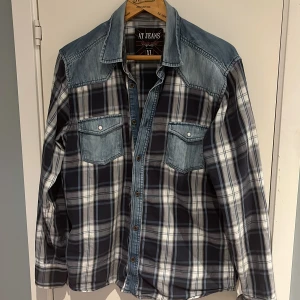 Rutig skjorta med denimdetaljer från AT JEANS - Säljer en snygg rutig skjorta från AT JEANS med ljusblå denim på axlar och bröstfickor. Skjortan har tryckknappar och två bröstfickor med lock. Perfekt för en avslappnad stil.