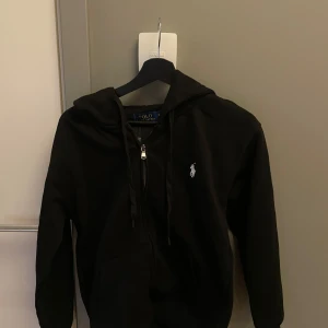 Svart zip hoodie från Polo Ralph Lauren - Säljer en svart zip hoodie från Polo Ralph Lauren med dragkedja och klassisk logga broderad på bröstet. Tröjan har huva med snörning och fickor framtill. SPRILLANS NY. Endast testad och det var fel storlek. Det är en S men funkar för S/Xs. Hör av dig vid eventuella frågor!🙌