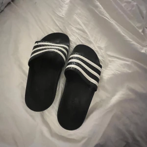 Svarta sandaler med vita ränder från Adidas - Säljer ett par klassiska svarta Adidas sandaler med tre vita ränder över foten. Perfekta att glida runt i hemma eller på stranden. Enkla att ta på och av, med bekväm passform och platt sula.(en liten fläck på vardera toffla)