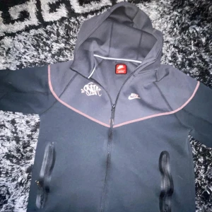 Svart Nike tech cyna world hoodiejacka med dragkedja och huva - Säljer en svart kofta från Nike med dragkedja och huva. Jackan har röda detaljer, två fickor med vattentäta dragkedjor och tryck på bröstet samt texten 'LIFEOFSYN' luvan. Perfekt för dig som gillar streetwear. (Byxor finns också) båda för 2000kr(nytt med kartongen på)