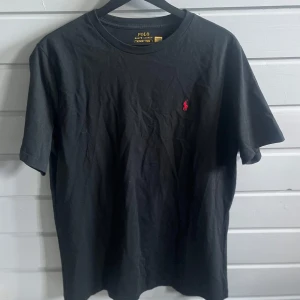 Svart t-shirt från Polo Ralph Lauren - Svart t-shirt från Polo Ralph Lauren med klassisk passform och liten röd broderad logga på bröstet. Perfekt basic-plagg med rund hals och korta ärmar. Enkel och stilren design.