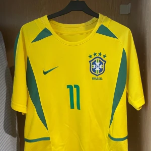 Brasilien fotbollströja Ronaldinho #11 - Säljer en retro Brasilien tröja med Ronaldinho 11. Bra skicka och inga defekter, perfekt för en stilren look till sommaren eller till träningen. Hör av dig om du undrar något eller kolla mina andra plagg!!✅🇧🇷