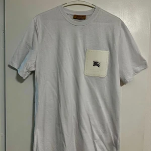 Vit t-shirt med ficka från burberry - Säljer en stilren vit t-shirt från burberry med en vit bröstficka som har en liten svart broderad detalj. T-shirten har normal passform och korta ärmar. Perfekt för en clean och enkel look.