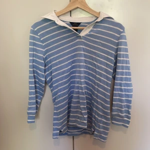 Blåvit randig pikétröja från Brooks Brothers - Säljer en blå och vit randig pikétröja från Brooks Brothers i 100% bomull. Tröjan har vit krage och trekvartsärm, perfekt för en avslappnad men ändå klassisk look. Snygg och tidlös design med mjukt material. Dock ett litet hål på vänsterarmen.