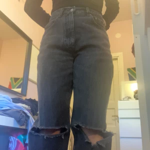 Gråsvarta jeans med hål från Gina Tricot - Säljer ett par svarta high waist jeans från Gina Tricot, modell 'perfect jeans'. Jeansen har raka ben och stora hål vid knäna för en trendig look. Klassisk femficksmodell med dragkedja och knapp.