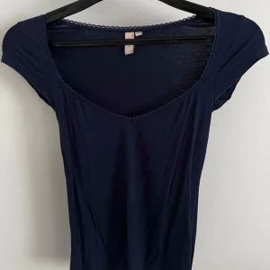 Mörkblå topp med spetskant - Säljer en mörkblå topp med v-ringning och fin spetskant längs halsringningen. Toppen har korta ärmar och är tillverkad i ett mjukt material som sitter skönt på kroppen. Perfekt till både jeans och kjol!