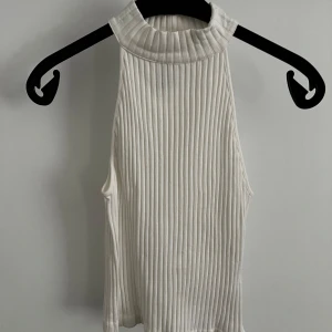 Vit ribbad ärmlös polotopp - Säljer en vit ribbad polotopp med hög krage och ärmlös design. Toppen har en mjuk och stretchig känsla, perfekt för att styla med jeans eller kjol. Enkel och stilren modell som passar till många olika looks.