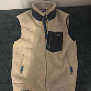 Patagonia väst - Säljer en beige fleeceväst från Patagonia med blå detaljer och svart bröstficka med dragkedja. Västen har två sidofickor med dragkedja och hög krage. Perfekt för lager-på-lager under kyliga dagar.