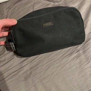 Svart necessär från Burberry - Säljer en stilren svart necessär från burberry med dragkedja upptill och rutigt handtag i ena änden. Perfekt för att organisera dina toalettartiklar när du är på resande fot. Klassisk design med Burberry-logga i metall framtill.