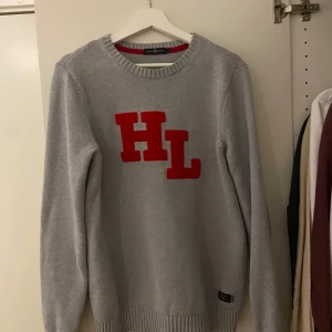 Grå stickad tröja med röda bokstäver HL - Säljer en grå stickad tröja från Henry Lloyd med stora röda bokstäver HL på bröstet. Tröjan har rund halsringning och långa ärmar med ribbade muddar vid ärmslut och nederkant. 