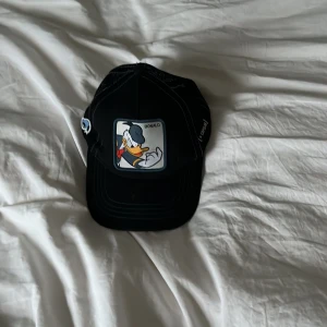Svart keps med Kalle Anka från Capslab - Svart keps med en stor patch framtill med Kalle Anka-motiv och texten 'Donald'. Kepsen har justerbar baksida i mesh och tydliga vita sömmar. Märkt med Capslab-logga på sidan.