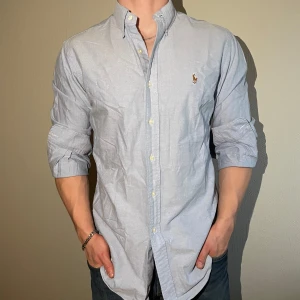 Ljusblå skjorta från Polo Ralph Lauren - Klassisk ljusblå skjorta från Polo Ralph Lauren med broderad logga på bröstet. Skjortan har lång ärm, knappar framtill och är i en custom fit-modell. Perfekt för en stilren och avslappnad look.