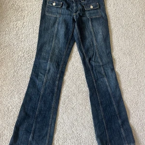 Blå bootcut jeans från H&M - Säljer ett par blå bootcut jeans från H&M med låg midja och justerbar vidd. Jeansen har framfickor med lock och knappar, klassisk denimlook och markerade sömmar. Perfekta för dig som gillar en retrostil!