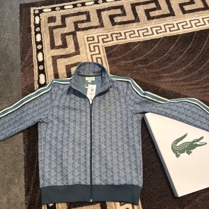 Blå mönstrad track jacket från Lacoste köpt för 2300 - Säljer en blå track jacket från Lacoste med vitt geometriskt mönster och klassisk krokodillogga på bröstet. Jackan har dragkedja, ribbade muddar och randiga detaljer längs ärmarna. Perfekt för en sportig och stilren look.