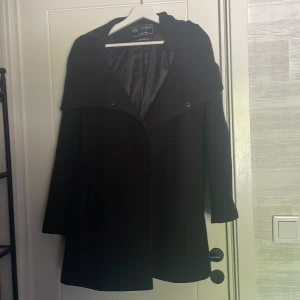 Svart kappa med huva från Amisu - Säljer en svart kappa från Amisu Classic Collection i storlek 44 med snygg huva och knäppning framtill. Kappan har ett klassiskt snitt och är helfodrad med diskret mönstrat innerfoder. Perfekt för kyligare dagar.