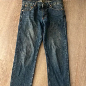 Mörkblå jeansbyxor från Woodbird - Klassiska mörkblå jeans från Woodbird med raka ben och fem fickor. Byxorna har orangea sömmar och knappgylf. Perfekta för dig som gillar en tidlös jeansstil.