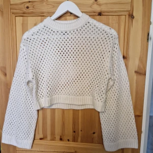 Vit croppad stickad tröja från H&M - Säljer en vit croppad stickad tröja från H&M. Tröjan har ett hålmönster över hela, ribbade muddar och lång ärm. Perfekt för lager-på-lager och ger en trendig look.