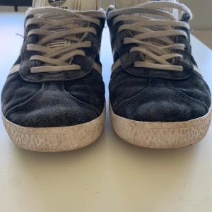 Marinblå Adidas Gazelle sneakers - Säljer ett par klassiska blå Adidas Gazelle sneakers med vita ränder och vit sula. Skorna har snörning och är tillverkade i mocka med detaljer i syntet. Perfekta för en sportig och stilren look.-AI. Nypris: 1079
