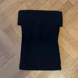 Svart tubtopp från Zara - Säljer en svart tubtopp från Zara i storlek XS. Toppen har en bred kant upptill och en snygg dragkedja längs sidan som detalj. Perfekt att styla till olika outfits!
