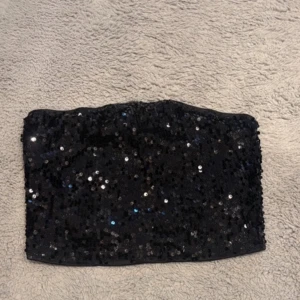 Svart paljett-tubtopp - Säljer en svart tubtopp täckt med glittrande paljetter. Perfekt för att sticka ut och ge en festlig känsla till outfiten. Toppens material är mjukt och stretchigt för en bekväm passform.