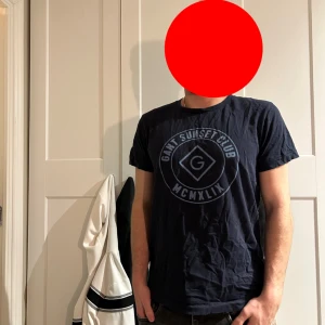 Mörkblå t-shirt från Gant - Säljer en mörkblå t-shirt från Gant med rund hals och tryckt logga på bröstet. T-shirten har normal passform och är kortärmad, perfekt för en avslappnad stil.