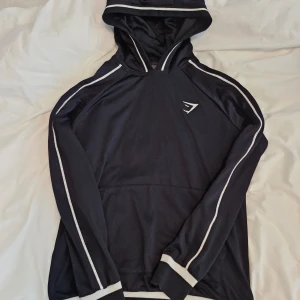 Sommar25. Svart hoodie från Gymshark med vita detaljer - Säljer en svart hoodie från Gymshark med vita ränder längs ärmarna och runt huvan. Stl S. Tröjan har lång ärm, huva och en klassisk sportig look. Perfekt för träning eller chill dagar.