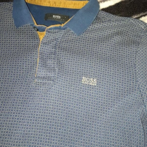 Blå mönstrad pikétröja från BOSS - Säljer en blå pikétröja från BOSS med små cirkelmönster och kontrastfärgad krage samt knappslå i gult. Tröjan har korta ärmar och broderad BOSS-logga på bröstet. Perfekt för en stilren och avslappnad look.