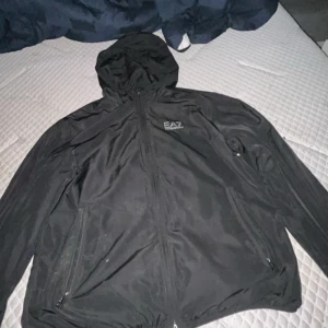 Svart vindjacka med huva från EA7 - Svart vindjacka från EA7 med huva och dragkedja framtill. Jackan har två sidofickor med dragkedja och EA7-logga på bröstet. Perfekt för blåsiga dagar och enkel att matcha med olika outfits. Sälj pga att jag ej använder dessa kläder längre