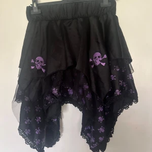 Svart kjol med lila dödskallemönster - Säljer en svart kjol med asymmetrisk skärning och lila dödskallar. Kjolen har volang och spetsdetaljer längs kanten, samt resår i midjan för extra komfort. Perfekt för dig som gillar alternativ stil! Helt ny. Helresår i midjan, onesize.