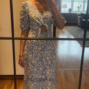 Blå blommig klänning med knyt och puffärm - Säljer en blå klänning med vita blommor, v-ringning och knyt framtill. Klänningen har kort puffärm, knappar längs hela framsidan och en liten öppning vid midjan. Perfekt för sommarens alla tillfällen!