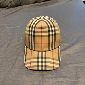Beige rutig keps från Burberry - Snygg keps från Burberry i klassiskt beige rutigt mönster med svarta, vita och röda linjer. Justerbar baktill med svart spänne och tydlig Burberry-logga. Väldigt få använd , bra skick!! Kepsen är köpt på farfetch kom dm för mer info och äkthetsbevis! Påse tillkommer