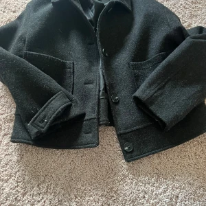 Svart kappa - Snygg svart kappa med knappar framtill och två stora fickor. Jackan har en klassisk krage och långa ärmar med dekorativa detaljer vid ärmsluten. Perfekt för kyligare dagar. Ena knappen på armen e borta men har fixat me en säkerhetsnål så enligt mig inget man tänker på alls. Kappan är i storlek M men passar absolut S 