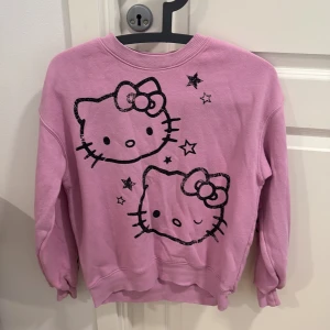 Rosa Hello Kitty tjocktröja - Säljer en söt rosa sweatshirt med Hello Kitty-tryck och stjärnor på framsidan. Tröjan har långa ärmar med texten 'HELLO KITTY' längs ena ärmen och stjärnor på den andra. Perfekt för alla Hello Kitty-fans!