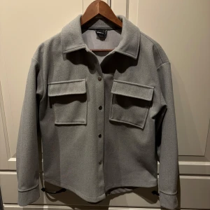 Grå kappa - Snygg, tunn grå kappa från Gina Tricot med knappar framtill och två stora bröstfickor. Perfekt för våren och sommaren! I storlek xs men passar även mig som är en S/M. Använd 1gång så inga defekter och i väldigt bra skick. Inga nopprar eller liknande!💕